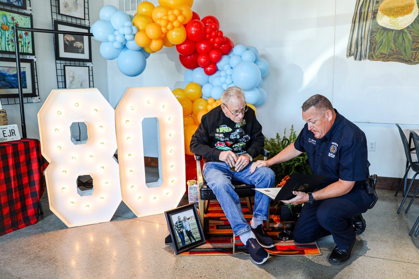 A firefighter kneels beside Buddy to present the mayor's letter, next to a light-up 80